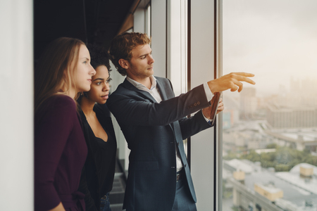 Handsome serious businessman in formal suite showing using digital tablet to his female colleagues plan of future site development from window of office located on high floor of modern skyscraperの写真素材