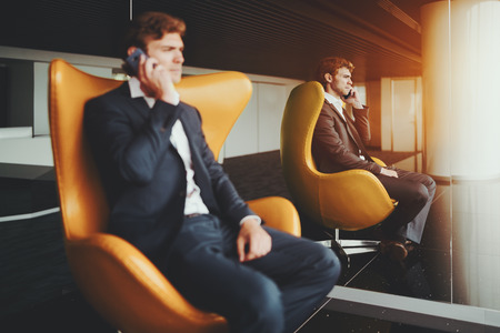 Serious bearded employer talking with his business colleague via smartphone while sitting on curved armchair, handsome businessman in formal suite having cell phone conversation in office interiorの写真素材