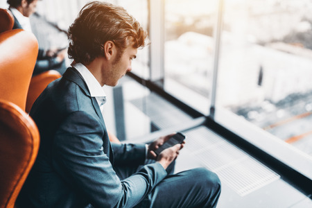 Adult handsome bearded businessman messaging via his smartphone with his colleagues, man entrepreneur in formal suite chatting on cellphone while sitting on armchair next to skyscraper office windowの写真素材