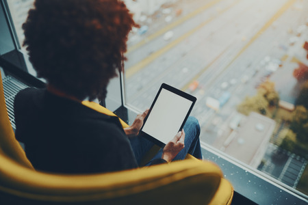 White mock-up of digital tablet screen in hands of black girl with afro hair sitting near window of skyscraper, blank template of digital pad screen in female hands of woman sitting in yellow armchairの写真素材