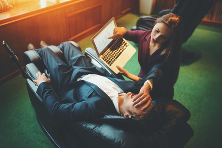 Scene from office life: businesswoman is showing her male boss information on screen of laptop but her colleague is chilling in black leather reclining massage chair and covering his face with palmの写真素材