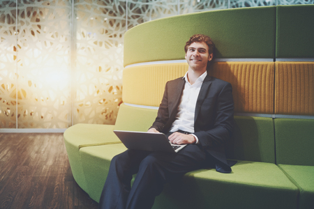 Handsome attractive smiling businessman is sitting with laptop on green and yellow curved sofa in modern meeting room with patterned glass wall behind and copy space for logo, your advert or messageの写真素材