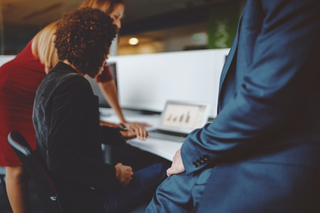 View of male hand of businessman in blue formal suit in office settings with two of his colleagues in defocused background working on laptop with graph on screen, shallow depth of fieldの写真素材