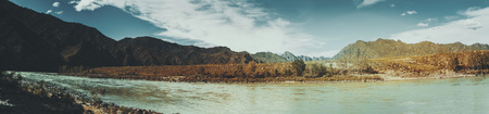 Panorama of Katun river from stony coastline, near Kuyus district, Altai mountains, Russia: shore with grass, rocks, birch and pine trees, fast water current, hills in distance, teal skyの写真素材