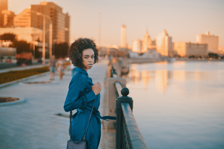 Charming young black tourist girl with afro hair, clutch and in blue dress standing near metal fencing of embankment of Moscow river during evening promenade with copy space zone for message or textの写真素材