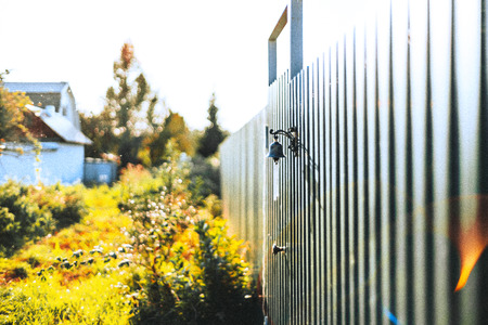 Brass little bell on metal striped fence of summer cottage stretching into distance with autumn greenery and other houses in blurred background, warm sunny day, Russiaの写真素材