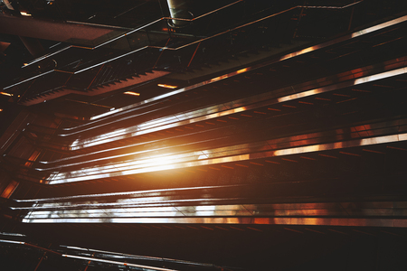 Wide-angle shooting of moving staircase in dark hall of modern airport terminal; interior of mall or railway station with empty escalators following opposite sides, strong flare from sun reflectionの写真素材