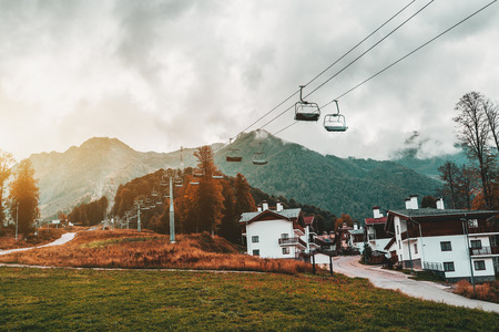 Autumn landscape: cableway stretching into distance over hills with houses of resort village, trees, cell connection towers, meadows and the road; low clouds, Estosadok district, Sochi, Russiaの写真素材