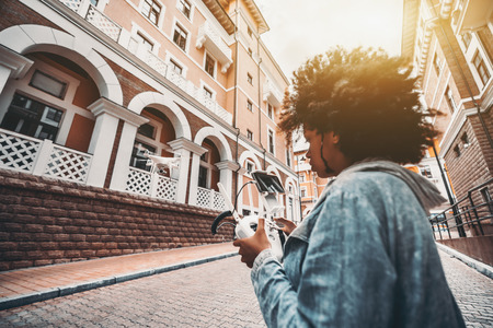 Flying drone in urban settings and black young woman with big curly hair operating this quadrupter using remote control device with joystick and connected smartphone as camera screen, sunny dayの写真素材