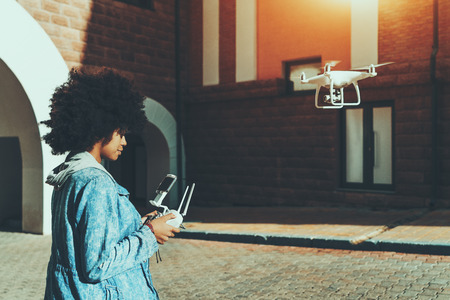 Young beautiful African American woman standing on pavement in urban settings and remotely tuning...