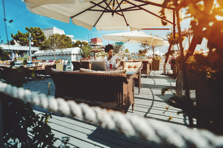 Wide-angle view of seaside cafe with beautiful young black lady sitting in at the wicker table in a distance with laptop, glass of red fruit drink, she is answering on the message in her smartphoneの写真素材