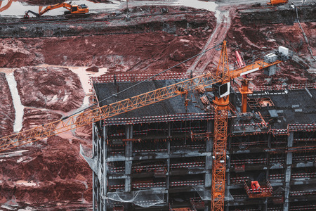 Orange modern crane lifting construction cargo to unfinished house on dull moody day in a huge pit full of reddish dirt and other construction machinery such as excavators, generators, cement feederの写真素材