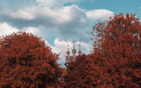 Carved wooden orthodox church cupola with crosses partly closed by trees of vivid unnatural reddish colors in foreground; cloudy sky in the background, overcast summer day, Moscow, Russiaの写真素材