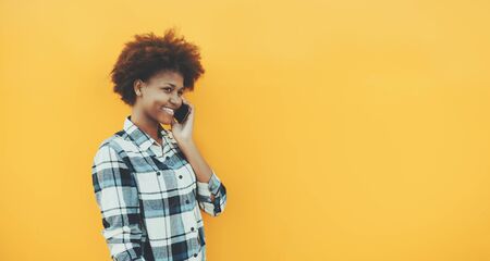 Cheerful young black female with perfect white smile is talking on the smartphone while standing in front of yellow solid wall; isolated portrait of smiling cute teenage student girl with cell phoneの写真素材