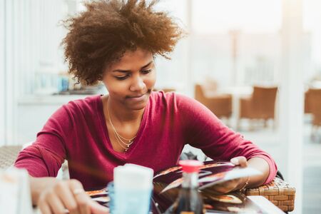Focused young Brazilian curly lady is sitting in street cafe outdoor, riffling pages of sushi menu booklet trying to choose delicious dish for herself; empty wicker armchairs in defocused backgroundの写真素材