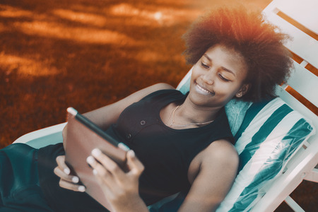 Cheerful Brazilian female laying on a deck chair and watching movie on her digital tablet; young black girl with afro laying hair on recliner and messaging via digital pad; vivid colors in backgroundの写真素材