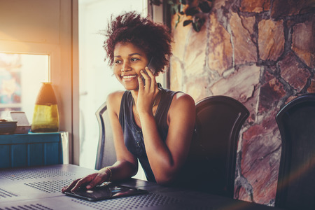 Smiling attractive young African American girl with curly hair is sitting in dark room of terrace and having phone conversation with her friend; tablet pc on the table, window with flare in backgroundの写真素材