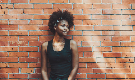Portrait of young curly charming black female standing in front of brick wall of summer house; sweet cute teenage African American girl next to brick surface with daisies in her curly Afro hairの写真素材