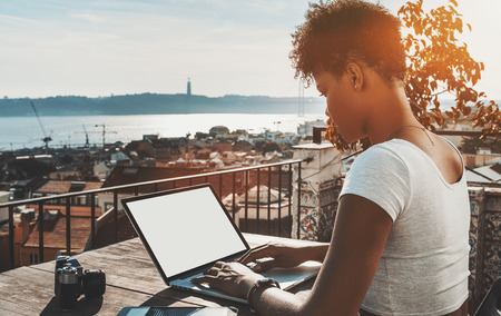 Side view of curly African American female copying recently made photos to laptop with blank mock-up screen while sitting on the balcony of Lisbon city with horizon; cityscape and river in distanceの写真素材