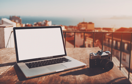 Modern laptop with empty white screen mock-up on the wooden table with retro film camera near; elegant netbook with empty screen template on table of cozy balcony in Lisbon city with cityscape behindの写真素材