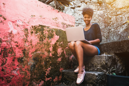 Young sweet smiling black female with laptop is sitting on stone step of staircase next to wall; cheerful curly Brazilian girl is sitting with netbook on old concrete staircase outdoorの写真素材