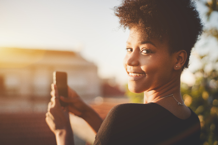 Close-up portrait of dazzling African American female with curly hair, partly turned to the camera while staying on the roof and taking pictures on mobile cell phone of cityscape on sunny summer dayの写真素材