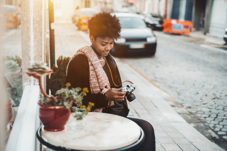 Exquisite young Brazilian tourist girl in coat and muffler is sitting at the marble table of street cafe next to sett paving and looking through photos on screen of her retro looking film cameraの写真素材