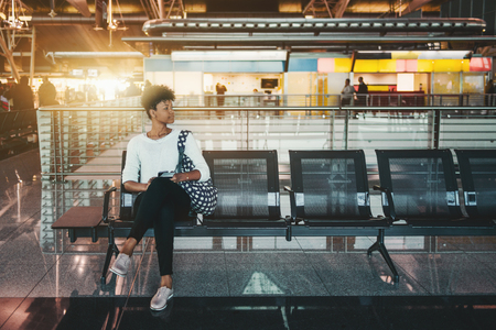 Lonely Brazilian girl is sitting inside of modern airport terminal on metal seats and waiting her flight; cute young African American female tourist is waiting for her train indoors of railway stationの写真素材