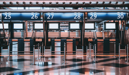 Front view of check-in area in contemporary airport: luggage accept terminals with baggage handling belt conveyor systems, multiple empty white informational LCD screen mockups, indexed check-in desksの写真素材