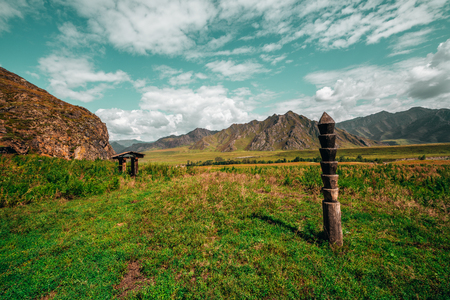 Wide-angle view of mountain scenery with old draw-well, wooden ancient sign as pillar, hills ridge in background, pasture with native grasses, valley, rocks, and teal skyの写真素材