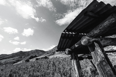 Wide-angle black and white view of old wooden abandoned draw-well in mountains with forsaken unfinished shacks in background, hill ridge, and meadow of cannabis and wormwood; copy space for your textの写真素材