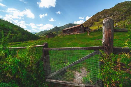 Wide-angle shooting of abandoned houses in forsaken village in mountains with pasture and wooden fence and Rabitz-type steel-wire plaster fabric in foreground; hill ridge in background, Altai, Russiaの写真素材