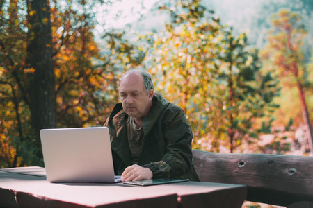 The mature confident businessman is working on his laptop with tablet pc near him on the table,...