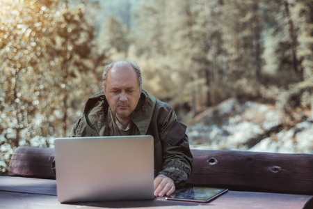 Portrait of balding mature businessman in coveralls with laptop and digital tablet sitting at wooden table during his vacation trip, with forest and hills in the defocused background; sunny autumn dayの写真素材