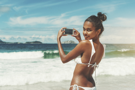 View from behind of sexy young black girl in swimsuit with wet skin, half-turned and looking at camera with smile while taking photos of ocean and horizon using her smartphone on sunny summer dayの写真素材
