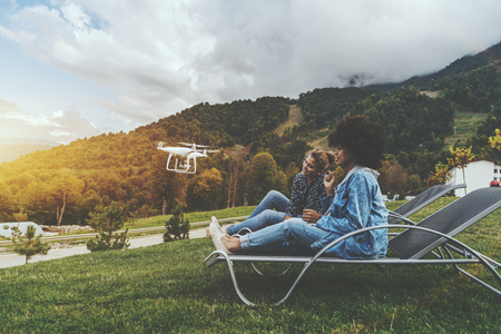 Two charming cheerful girls of different races on deck chairs on the lane are sitting to the camera of the flying drone while it filming them; with hill ridge in the background; autumn morningの写真素材