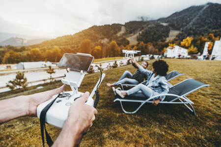 Male hands in foreground holding the remote controller for flying drone operation and two girls in the defocused background laying on deck chairs and touching this multicopter while it filming themの写真素材