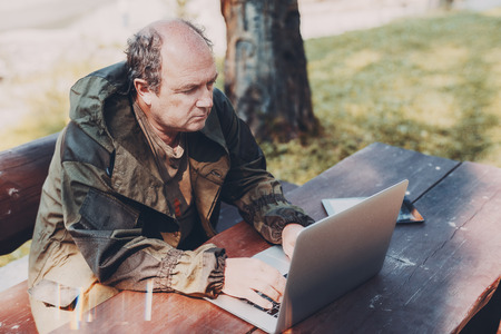 Aged partly bald man is sitting in a park with the laptop and digital tablet; adult male gamekeeper is sitting at the wooden table outdoors next to the tree and working on his netbook on a sunny dayの写真素材