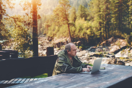 Elderly man employer is spending his vacations with work outdoors: sitting with the laptop at the wooden table with a digital tablet next to him and summer pine forest in a defocused backgroundの写真素材