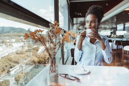 Charming young African American lady is sitting inside of a restaurant on the top-floor of a building, holding the cup of a hot delicious tea and thoughtfully looking out the window on a cityscapeの写真素材