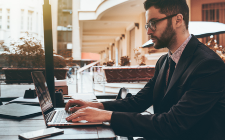 Side view of a serious bearded man entrepreneur sitting with laptop outdoors in a bar and doing remote work; handsome businessman in glasses in street cafe with his netbook preparing for the meetingの写真素材