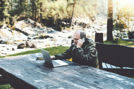Elderly businessman with the laptop in a dark-green overalls having a conversation via smartphone while sitting outdoors in the forest at the wooden table, working remotely during his vacationsの写真素材