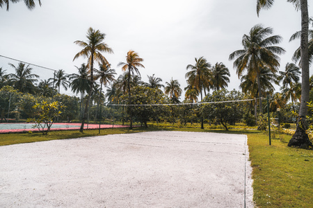Side wide-angle view of the volleyball court: coral sand on the ground, multiple palm trees and other plants around, paled tennis court on the right; bright summer day in a luxury Maldives resortの写真素材