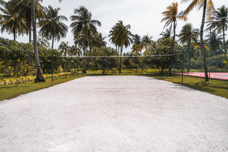 Wide-angle frontal view of the volleyball court: coral sand on the ground, multiple palm trees and other plants around, paled tennis court on the right; bright summer day in the Maldives resortの写真素材