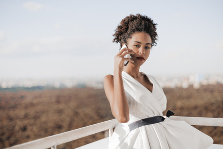Serious charming African American lady in a white dress waving in the wind is speaking on the smartphone while standing on an observation balcony of a skyscraper with the defocused cityscape behindの写真素材