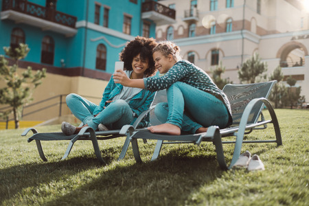 Happy interracial female couple: smiling Caucasian girl is showing photo on the screen of the smartphone to her black laughing friend while both sitting on the recliners on the spring gladeの写真素材