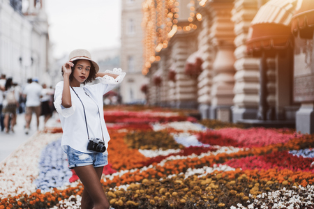 Young exquisite African-American girl with the winsome smile in the hat and with retro film photo camera standing on a street in urban settings with flowerbed behind and copy space area on the rightの写真素材