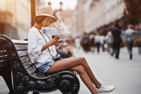Dainty African-American female in a white chemise and hat and with retro film photo camera is sitting outdoors on a wooden bench of a busy evening street and having chat with family via the smartphoneの写真素材