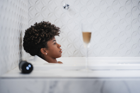 Side view of a tired young curly black girl laying, relaxing in the bath indoors and listening to the music form a wireless speaker next to her and a glass of a white wine in the foregroundの写真素材