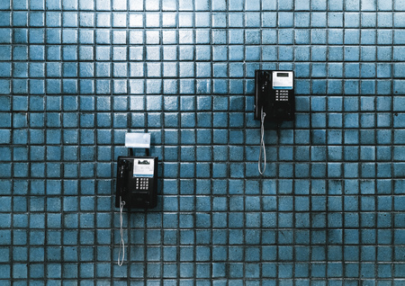 Two black public telephones on a teal wall of ceramic tiles indoors at rail station depot; two...